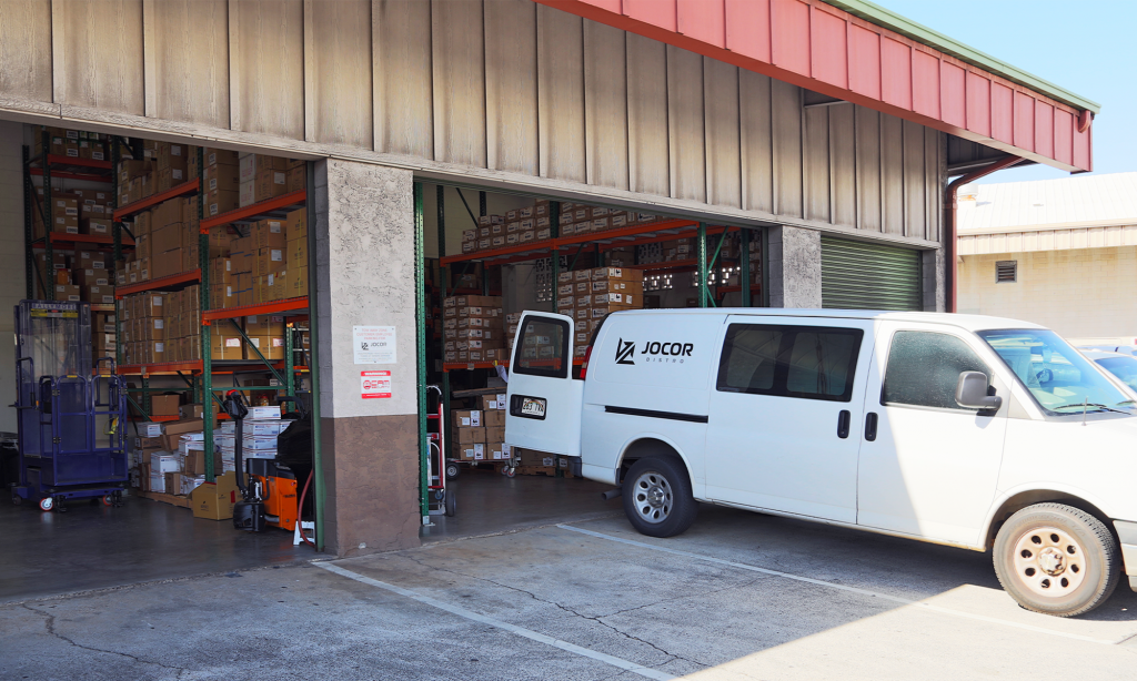 Outside of JOCOR Distro Warehouse with rolling gates open and JOCOR Distro cargo van being filled for delivery.